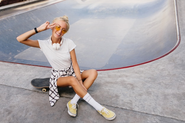 Lustige Skaterfrau mit trendiger Frisur, die positive Emotionen ausdrückt. Außenporträt des freudigen weiblichen Modells, das im Skatepark kühlt.