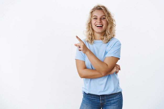 Lustige und fröhliche, optimistische europäische Frau der 20er Jahre mit kurzen lockigen blonden Haaren und blauen Augen, die aufrichtig und glücklich lächelt, als sie auf die obere linke Ecke auf das Produkt zeigt, das sie mag, und breit in die Kamera lächelt