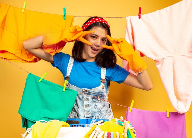 Lustige und schöne Hausfrau, die Hausarbeit lokalisiert auf gelbem Hintergrund tut. Junge kaukasische Frau, die durch gewaschene Kleidung umgeben ist. Häusliches Leben, helle Kunstwerke, Haushaltskonzept. Posieren, lächelt.