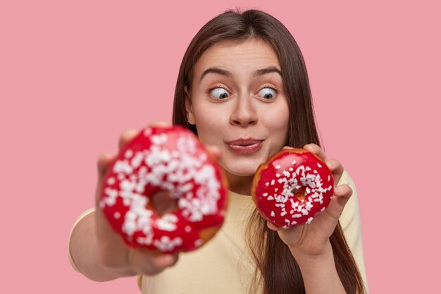 Lustige Zeit mit Essen. Stilvolle überraschte brünette Frau isst köstliche Donuts, sieht mit Erstaunen und Spaß aus