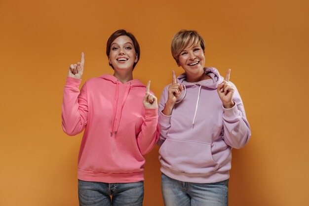 Lustige zwei Damen mit kurzer kühler Frisur in den modernen rosa Kapuzenpullis und in den Jeans, die Daumen oben auf orange isoliertem Hintergrund zeigen.
