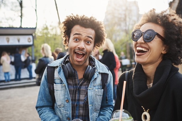 Lustiger emotionaler junger dunkelhäutiger Mann mit Afro-Haarschnitt, der Gesicht an der Kamera macht, während Zeit im Park mit bester Freundin verbringt, Tee trinkt und schönen Abend genießt.