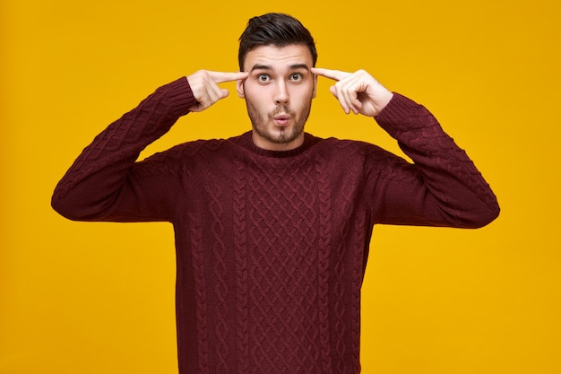 Lustiger emotionaler junger Mann im kuscheligen kastanienbraunen Pullover, der schockierten Blick hat und beide Vorderfinger an seinen Schläfen rollt,