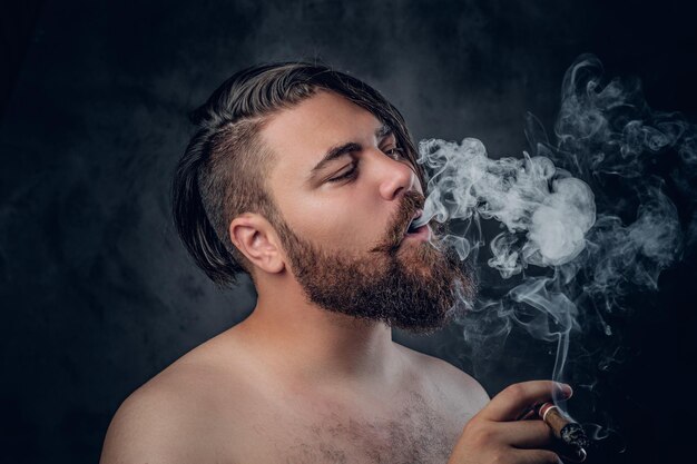 Lustiger hemdloser bärtiger Mann, der eine Zigarre auf grauem Hintergrund raucht.