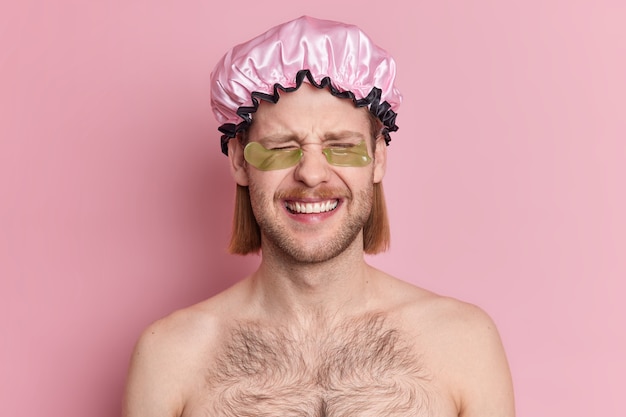 Lustiger hemdloser junger Mann trägt Badehut und Hydrogelflecken unter den Augen genießt Schönheitsbehandlungen nach dem Duschenlächeln glücklich hat Borsten- und Bobfrisur.
