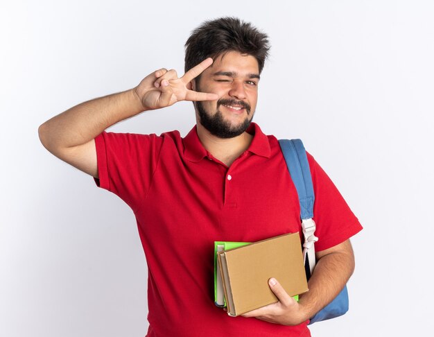 Lustiger junger bärtiger Student im roten Poloshirt mit Rucksack, der Notizbücher hält und lächelnd und zwinkernd mit V-Schild stehend aussieht looking