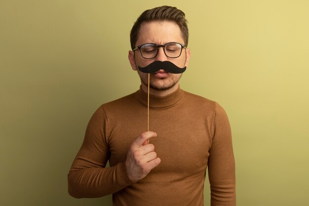 Lustiger junger blonder gutaussehender Mann mit Brille, der einen falschen Schnurrbart auf einem Stock über den Lippen hält und auf den Schnurrbart schaut, der auf der olivgrünen Wand isoliert ist?