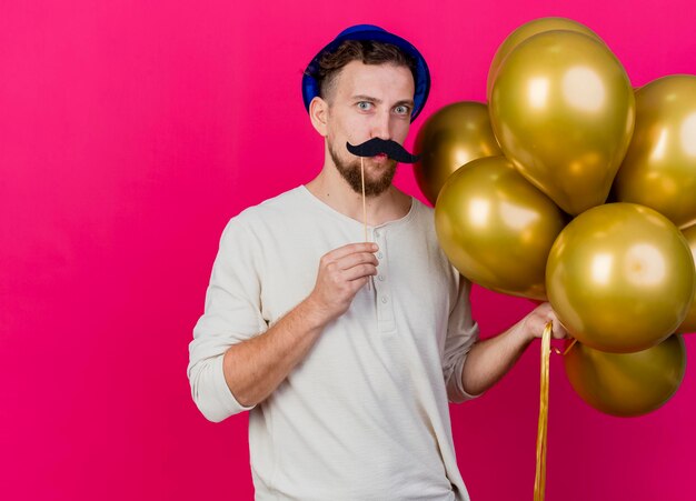 Lustiger junger hübscher slawischer Party-Typ, der Partyhut hält, der Luftballons und falschen Schnurrbart auf Stock über Lippen hält, die Front betrachten, die Kussgeste lokalisiert auf rosa Wand mit Kopienraum tut