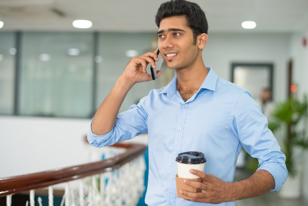 Lustiger junger männlicher Manager, der am Telefon spricht und Kaffee trinkt