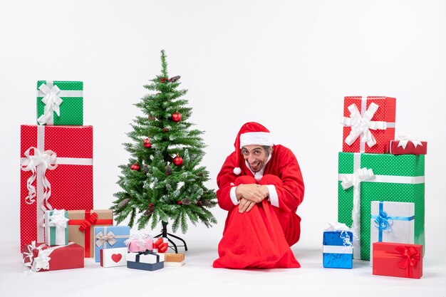 Lustiger junger Mann verkleidet als Weihnachtsmann mit Geschenken und geschmücktem Weihnachtsbaum, der auf dem Boden auf weißem Hintergrund sitzt