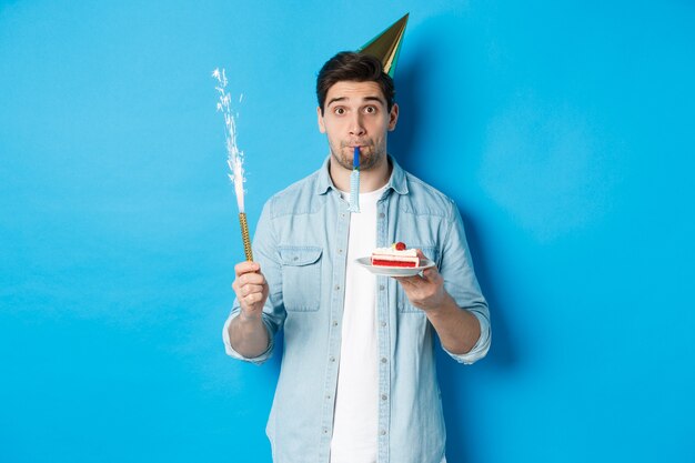 Lustiger Kerl feiert Geburtstag, hält B-Day-Kuchen, Feuerwerk und trägt Partyhut, steht auf blauem Hintergrund.
