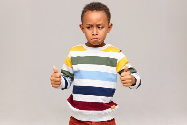 Lustiger kleiner afroamerikanischer Junge im gestreiften Pullover, der aufgibt, Daumen aufgibt, gut gemacht sagt, jemanden für guten ausgezeichneten Job, Erfolg beim Studium oder bei der Arbeit lobend. Schwarzes Kind genehmigt