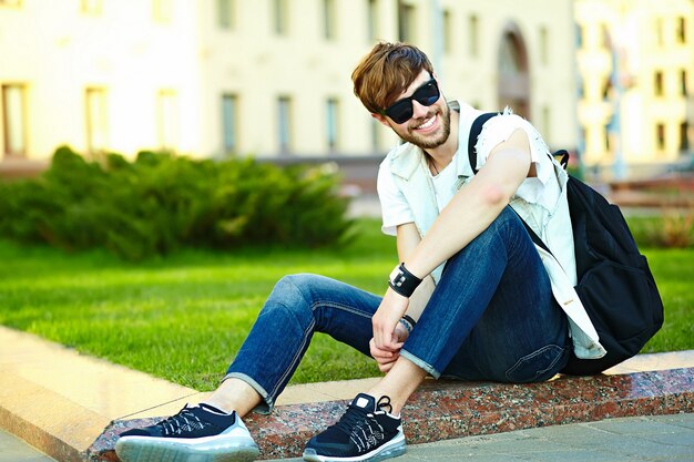 Lustiger lächelnder Kerl des gutaussehenden Mannes des Hippies im stilvollen Sommerstoff in der Straße, die auf Gras im Park sitzt