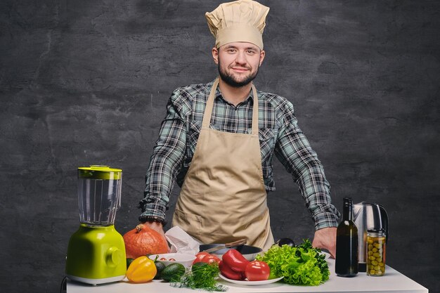 Lustiger männlicher Koch, der neben dem Tisch mit viel frischem Gemüse posiert.