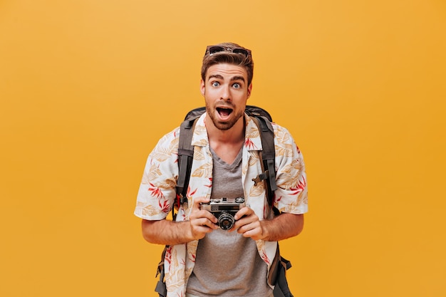 Lustiger Tourist mit blauen Augen in Sonnenbrille, modischem Sommerhemd und grauem kariertem T-Shirt mit Blick in die Kamera auf oranger Wand camera