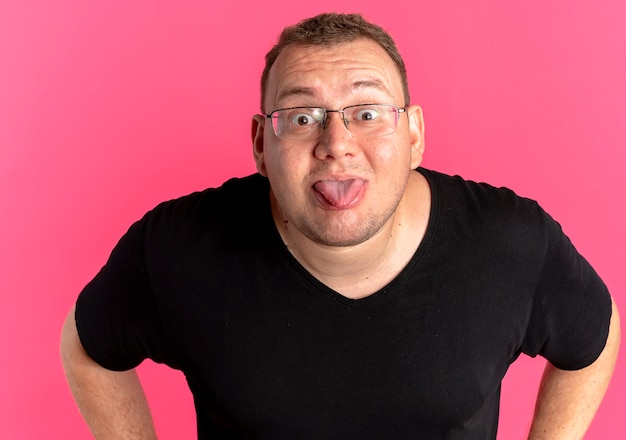 Lustiger übergewichtiger Mann in der Brille, die das schwarze T-Shirt trägt, das Zunge heraussteht, die über rosa Wand steht