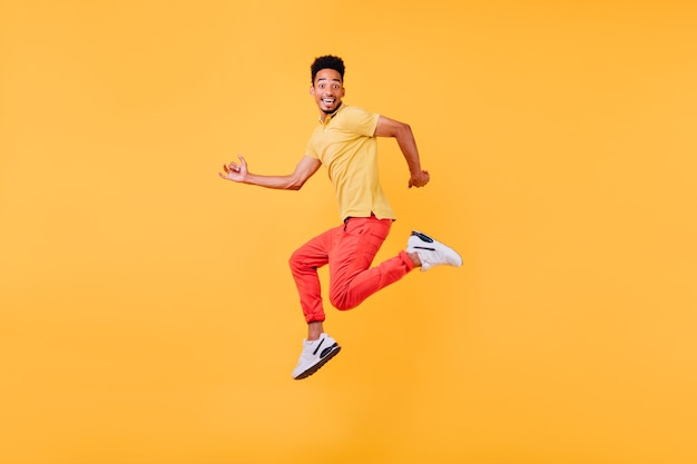 Lustiges afrikanisches männliches Modell, das mit überraschtem Lächeln aufwirft. Innenfoto des springenden sportlichen schwarzen Mannes.