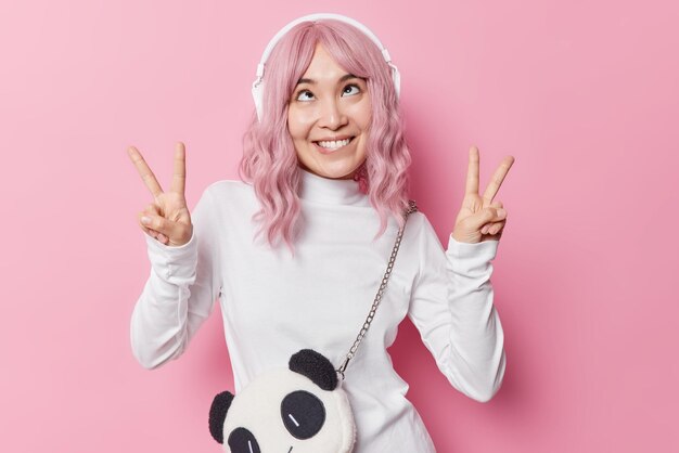 Lustiges asiatisches Mädchen macht Grimassen, zeigt Friedensgesten, täuscht herum, hört Lieblingslied, genießt Melodien, die in Kopfhörern gespielt werden, trägt lässigen weißen Rollkragenpullover, posiert als Zubehör für drahtlose Geräte im Innenbereich