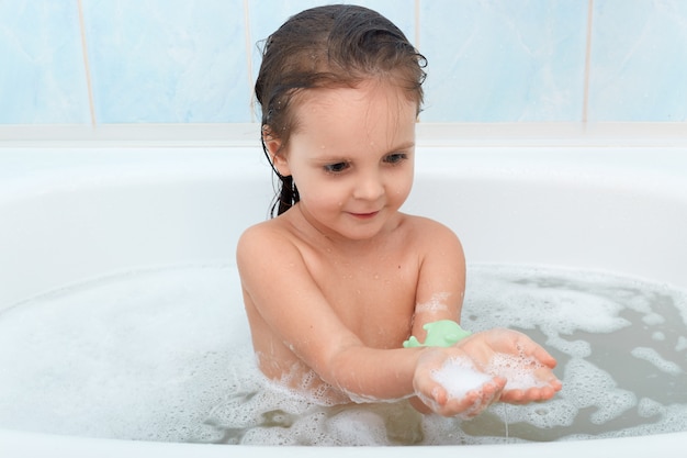 Lustiges Baby, das mit Wasser und Schaum in der großen Badewanne spielt