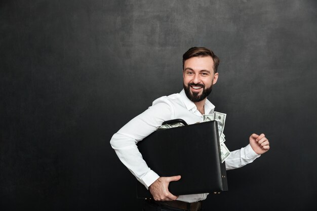 Lustiges Bild des glücklichen Mannes schwarzen Aktenkoffer voll halten von den Dollarbanknoten nach innen, lokalisiert über dunkelgrauem