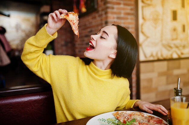 Lustiges brünettes Mädchen im gelben Pullover, das Pizza im Restaurant isst