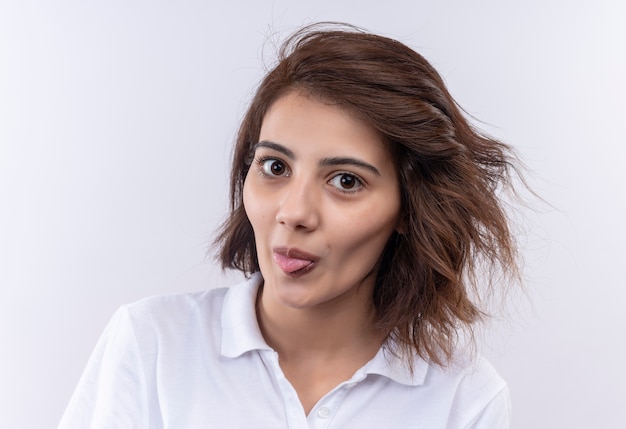 Lustiges fröhliches junges Mädchen mit kurzen Haaren, die weißes Poloshirt tragen, das Kamera betrachtet, die Zunge herausragt