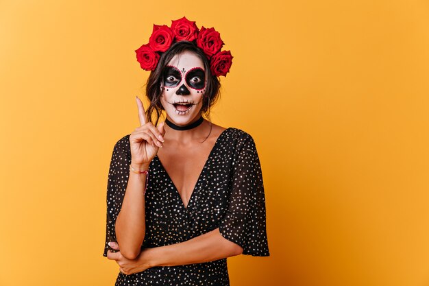 Lustiges gebräuntes Mädchen mit gemaltem Gesicht erinnerte sich an interessante Gedanken. Porträt der Frau mit Rosen auf dem Kopf im orangefarbenen Atelier.