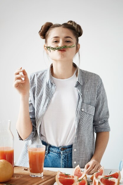 Lustiges junges Mädchen mit Brötchen, die Schnurrbart mit Rosmarinbrunch machen, der gesunden Grapefruit-Entgiftungs-Smoothie über weißer Wand vorbereitet.