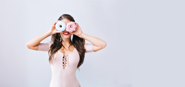 Lustiges junges Mädchen mit langen Haaren, die bunte Donuts gegen ihre Augen halten. Attraktiver Teenager, der Spaß mit Süßigkeiten hat, lecker. Heller Lebensstil.