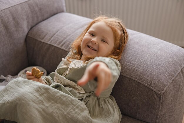 Lustiges kleines kaukasisches Mädchen mit roten Haaren lächelt und zeigt mit dem Finger auf die Kamera, während es auf der Couch liegt Kinderkonzept