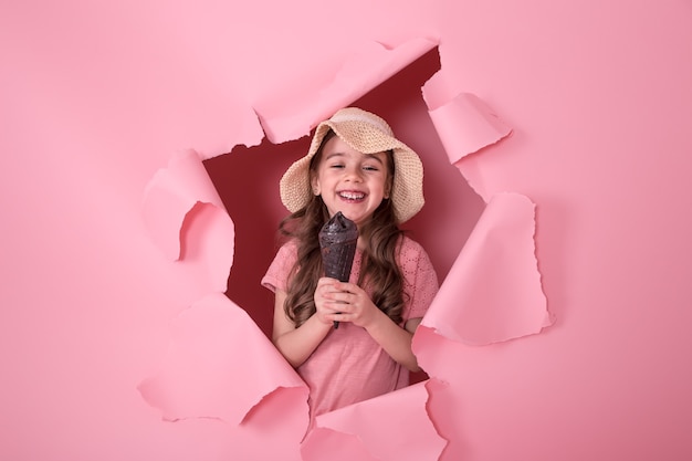 lustiges kleines Mädchen, das aus dem Loch in einem Strandhut und Eiscreme in ihren Händen späht, auf einem farbigen rosa Hintergrund, Raum für Text, Studio-Schießen