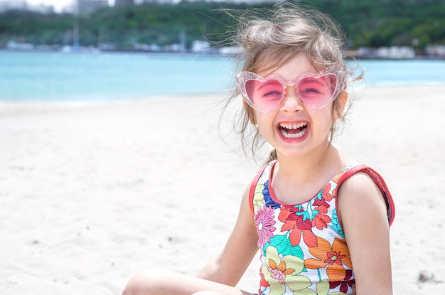 Lustiges kleines Mädchen, das in der Sonnenbrille aufwirft, die mit Sand am Strand spielt. Sommerunterhaltung und Erholung.