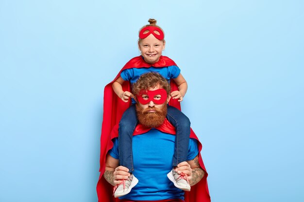 Lustiges kleines Mädchen im Superheldenkostüm, sitzt auf den Schultern des Vaters, lässt seine Ohren herausragen, hat Spaß mit Papa