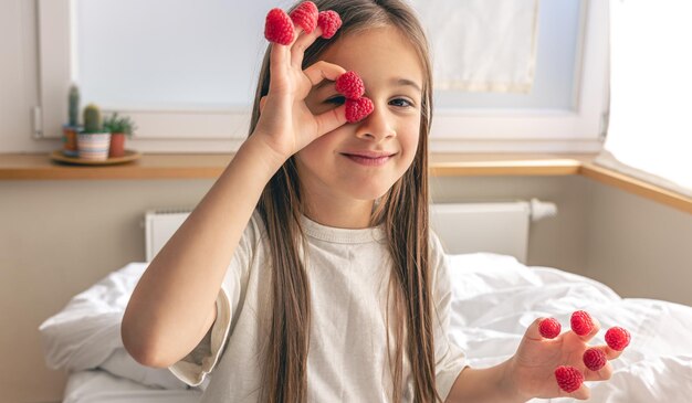 Lustiges kleines Mädchen mit Himbeeren an den Fingern morgens im Bett