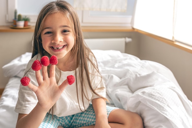 Lustiges kleines Mädchen mit Himbeeren an den Fingern morgens im Bett