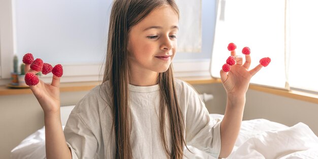 Lustiges kleines Mädchen mit Himbeeren an den Fingern morgens im Bett