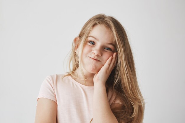 Lustiges kleines Mädchen mit schönen langen blonden Haaren, Wange mit der Hand drückend, lustige Gesichtsausdrücke machend, Spaß habend ..