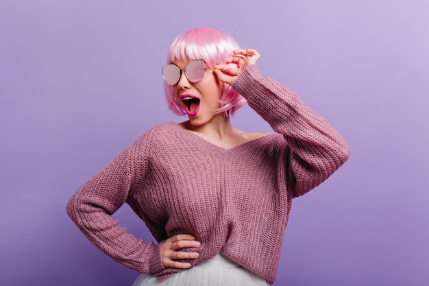 Lustiges mädchen im gestrickten pullover und im rosa peruke, die mit vergnügen tanzen. innenporträt der sorglosen dame in der perücke, die in der sonnenbrille aufwirft.