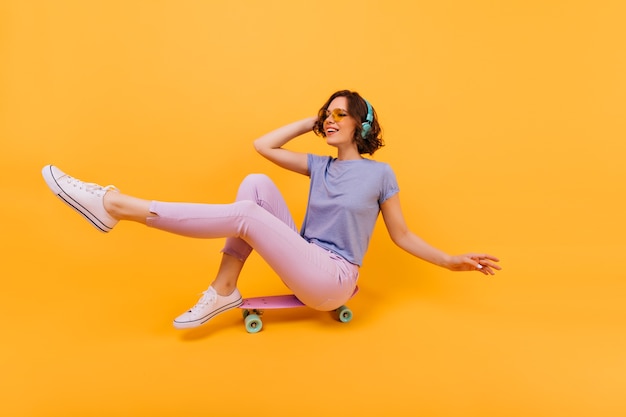 Lustiges Mädchen in der rosa Hose, die auf Skateboard sitzt und Gesichter macht. Innenfoto der reizenden weißen Frau in den Kopfhörern