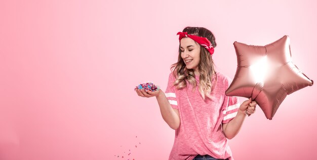 Lustiges Mädchen in einem rosa T-Shirt mit Luftballons und Konfetti gibt ein Lächeln und Emotionen auf einem rosa Hintergrund