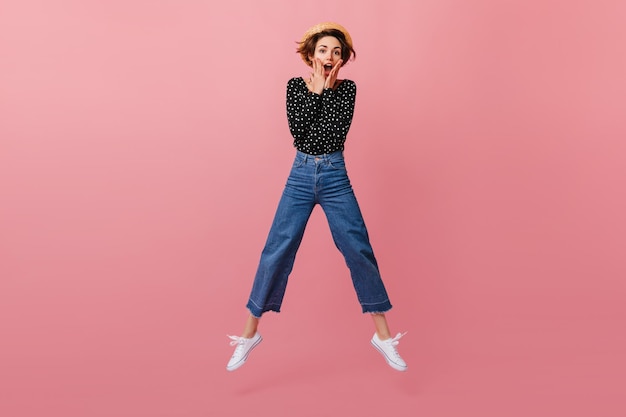 Lustiges Mädchen mit Strohhut, das auf rosa Hintergrund springt Studioaufnahme einer emotionalen Frau in Vintage-Jeans, die tanzt und in die Kamera schaut