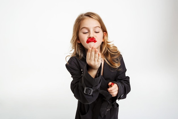 Lustiges Mädchen mit unordentlichem Make-up. Tragendes modisches schwarzes Trench-Luftküssen, hoding roten Lippenstift.