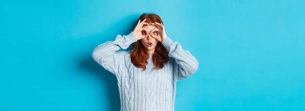 Kostenloses Foto lustiges rothaariges weibliches model im pullover, das durch die fingerbrille auf die kamera starrt und etwas int sieht