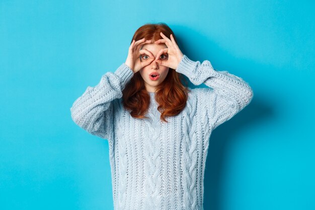 Lustiges rothaariges weibliches Modell im Pullover, das durch die Fingerbrille auf die Kamera starrt, etwas Interessantes sieht und auf blauem Hintergrund steht
