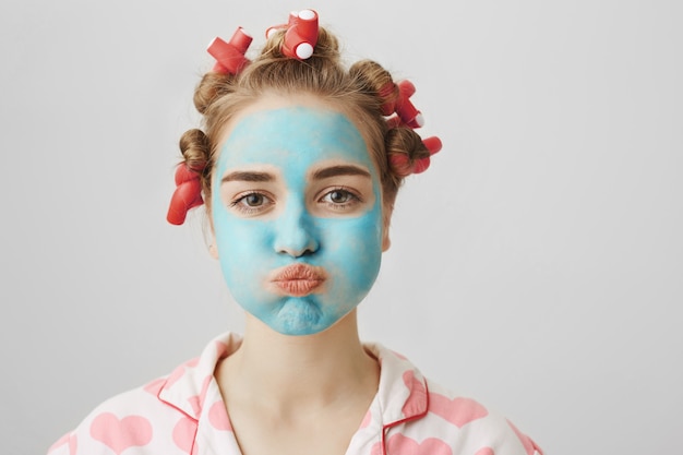 Lustiges süßes mädchen in den lockenwicklern und in der gesichtsmaske, die schmollen und atem halten