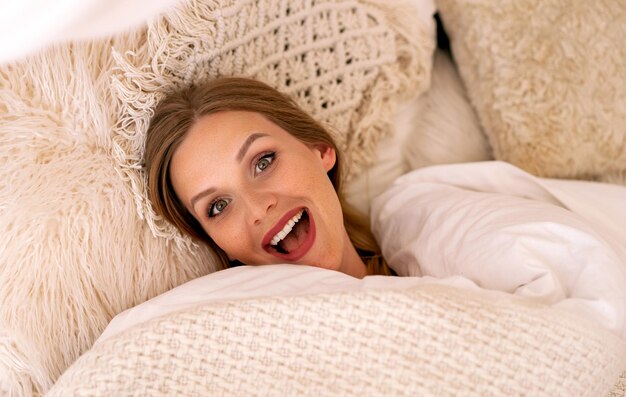 Lustiges süßes Morgenporträt einer blonden Frau, die gerade im Schlafzimmer im Boho-Stil aufgewacht ist Glückliches positives Gesicht