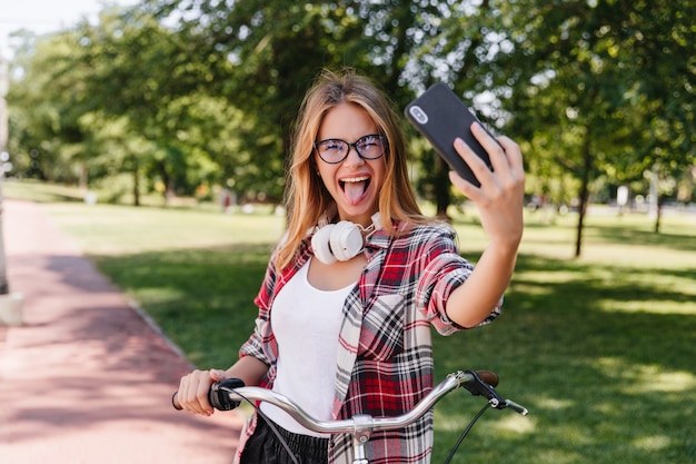 Lustiges weibliches Modell, das im Park mit Zunge heraus aufwirft. Außenporträt des aktiven Mädchens auf dem Fahrrad, das selfie mit Lächeln macht.
