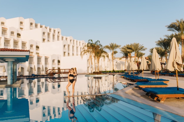 Luxuriöses weißes Hotel Ägypten im östlichen Stil, Resort mit schönem großen Pool. Hübsches Mädchen, Modell, das schwarzen Badeanzug trägt, der in der Mitte des Pools aufwirft. Urlaub, Ferien, Sommerzeit.