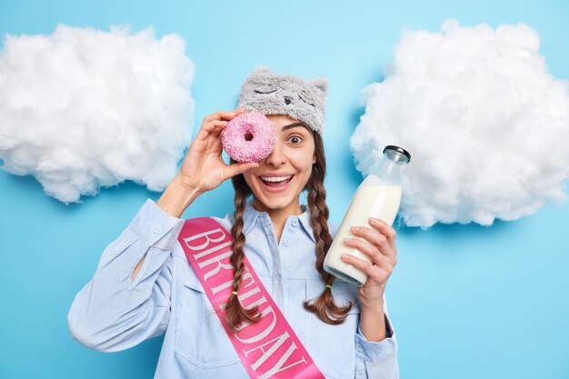 Mädchen cons Auge mit köstlichem glasiertem Donut auf dem Auge lächelt angenehm hält Glasflasche Milch feiert Geburtstag allein isoliert auf Blau