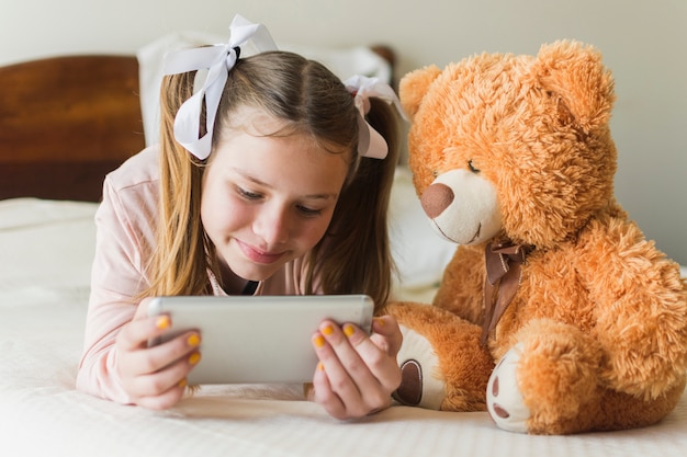 Mädchen, das auf Bett mit dem Teddybären betrachtet Handy liegt